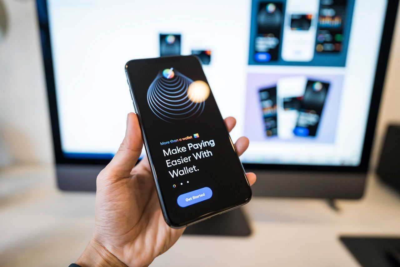 Main tenant a smartphone showing a digital wallet app interface, with a blurred monitor in the background.